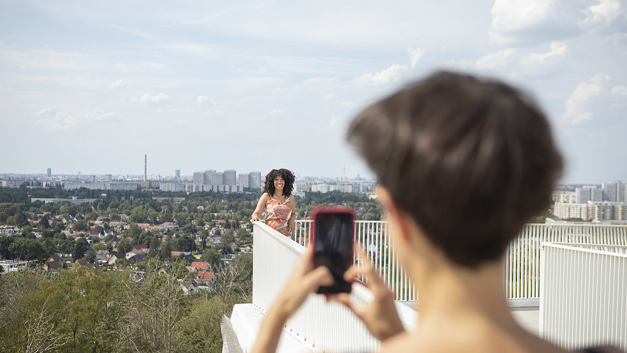 Eine Frau fotografiert mit einem Handy eine andere Frau auf dem Wolkenhain im Kienbergpark mit der Stadt im Hintergrund